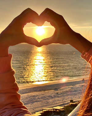 hands in a heart shape around the sun, facing the ocean coastline