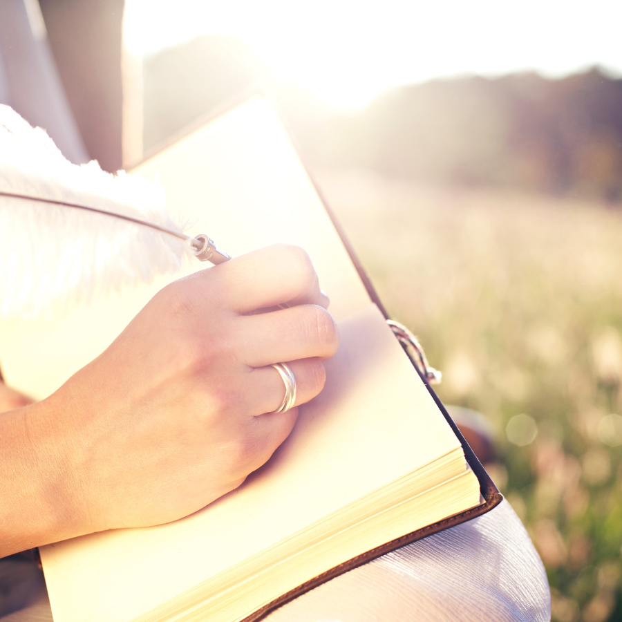 Person sitting outdoors, holding a journal and pen, writing in the sunlight