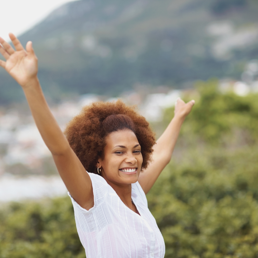 A woman smiling with hands raised in an outdoor setting, letting go of expectations and embracing imperfections for a stress-free holiday season