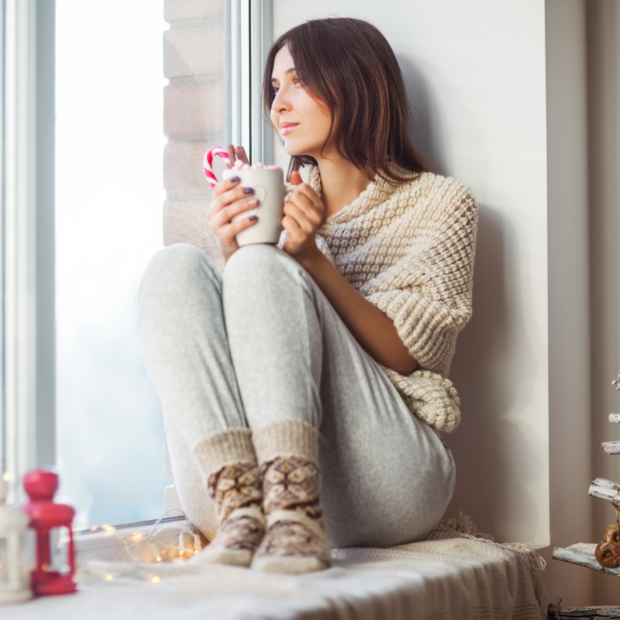 Woman sitting on a cozy window ledge in loungewear, enjoying a cup of coffee. Illustrates self-care during the holiday season for a stress-free experience.