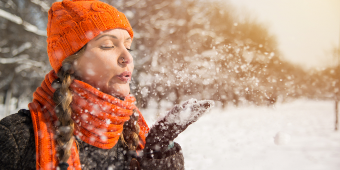 A woman standing in the snow, blowing snow out of her hand. She is wearing a warm coat, hat, and gloves, and appears to be enjoying the serene winter landscape. This image represents finding inner peace and stress relief during the holiday season with expert-approved self-care tips